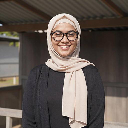 Photograph of a smiling woman wearing a white hijab, black cardigan, and glasses, standing outdoors under a wooden roof structure.