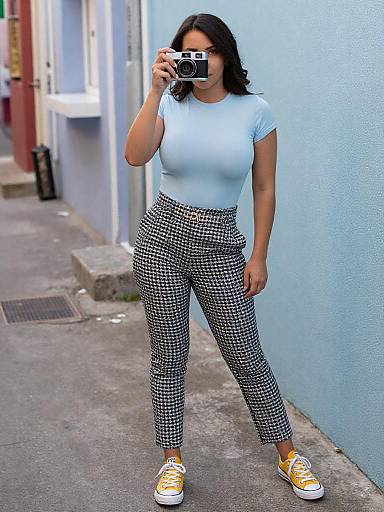 Photograph of a woman with medium skin tone, black wavy hair, wearing a white t-shirt, black-and-white checkered pants, and yellow