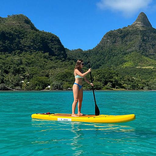 Photograph of a woman with dark hair in a white bikini, standing on a yellow paddleboard, paddling in turquoise water with lush green mountains in