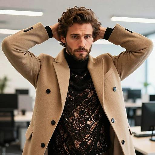 Photograph of a handsome, bearded man with curly brown hair, wearing a beige coat over a black lace turtleneck, hands behind head,
