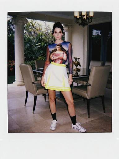 Photograph of a woman with dark hair, wearing a sheer black top, yellow and white skirt, black socks, and white sneakers, standing in a