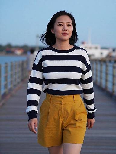 Photograph of an Asian woman with shoulder-length black hair, wearing a black-and-white striped sweater and mustard yellow shorts, standing on a wooden pier with