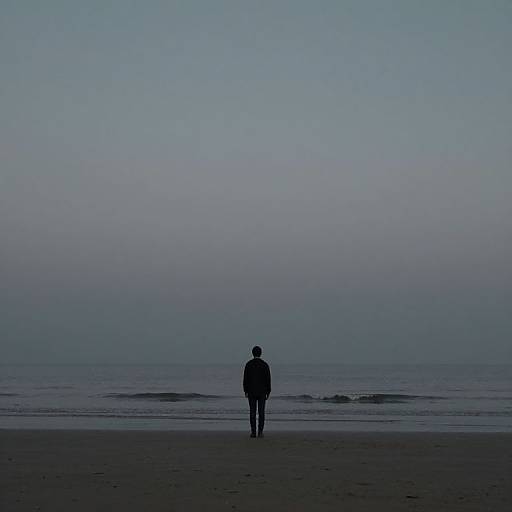 Photograph of a lone person, silhouetted in dark clothing, walking on a deserted beach at dusk, with a calm, blue-gray ocean