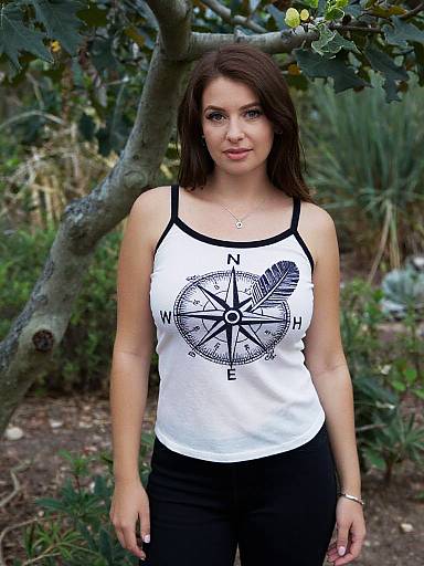 Photograph of a brunette woman with fair skin, wearing a white tank top with a compass design and black pants, standing outdoors by a tree in a