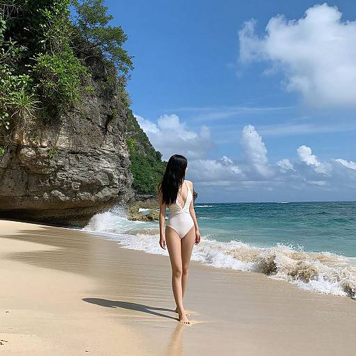 Photograph of a woman in a white one-piece swimsuit walking on a sunny, sandy beach with turquoise waves and a rocky, forested cliff in