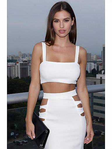 Photograph of a slender, brunette woman with long hair, wearing a white, cut-out crop top and high-waisted skirt, holding a black