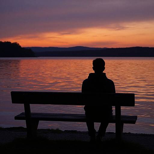 Silhouetted person sitting on a bench at sunset, reflecting orange and purple hues on a calm lake with distant hills.
