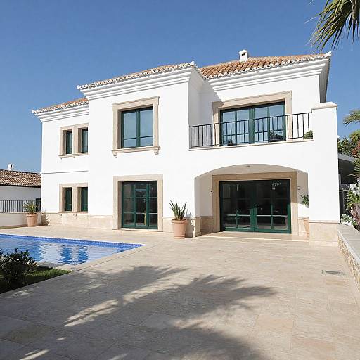 Photograph of a two-story white Mediterranean-style house with red-tiled roof, black-framed windows, balcony, pool, and clear blue sky.