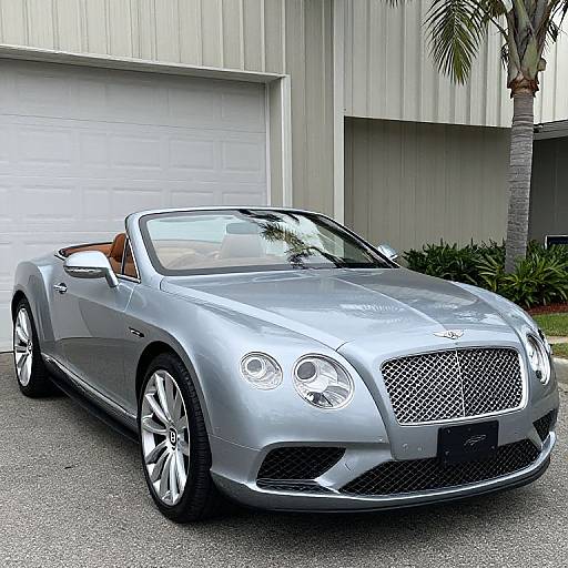 Photograph of a sleek, silver convertible Maserati with its top down, parked in front of a white garage door and modern building, with a