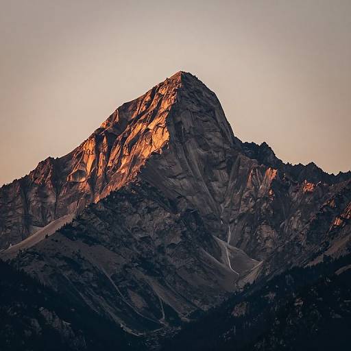 Photograph of a rugged mountain peak bathed in warm sunset light, with dark shadows and a clear, pale sky background.