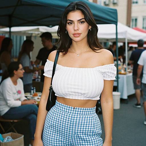 Photograph of a young woman with dark hair, wearing a white off-shoulder crop top and blue checkered skirt, standing in a bustling outdoor