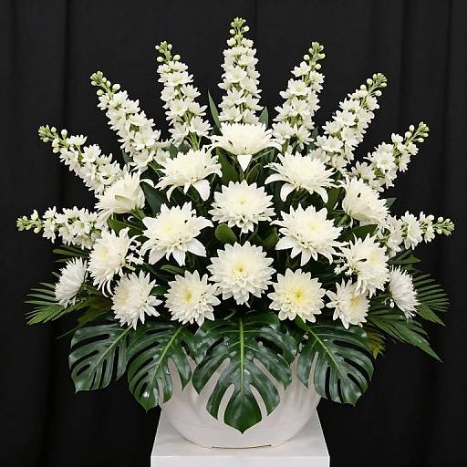 Photograph of a white floral arrangement featuring chrysanthemums, white snapdragons, and monstera leaves, set against a black backdrop