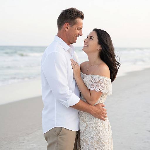 Photograph of a smiling couple embracing on a beach, the man in a white shirt and beige pants, the woman in an off-shoulder lace