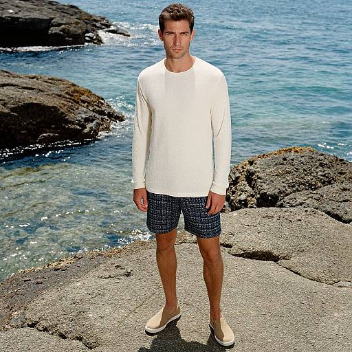 Photograph of a man with short brown hair, wearing a white long-sleeve shirt, navy blue patterned shorts, and beige slip-on shoes