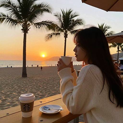 Photograph of a woman in a white knit sweater, holding a coffee cup, sitting at a beachside table, palms and sunset in background.