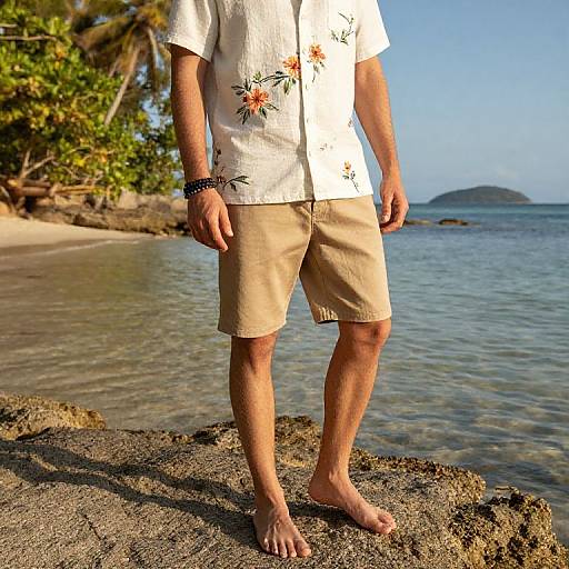 Photograph of a man's lower torso, wearing a white floral shirt, beige shorts, and a black bracelet, standing barefoot on a rocky beach