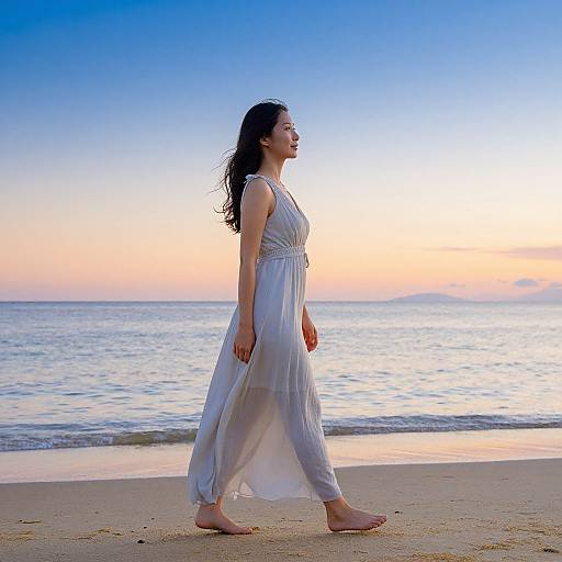 Photograph of a young woman with long black hair in a flowing white dress walking barefoot on a beach at sunset.