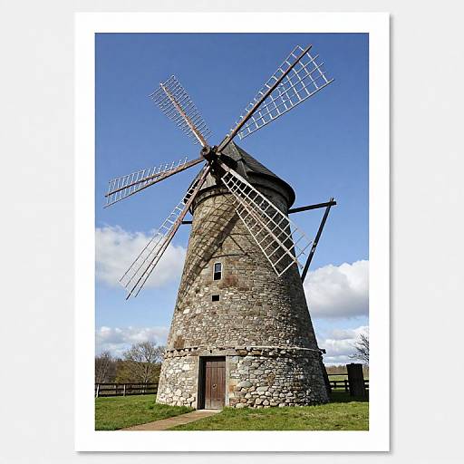 Photograph of a rustic, stone windmill with large, lattice-bladed sails, set against a bright blue, partly cloudy sky. Grass and fence