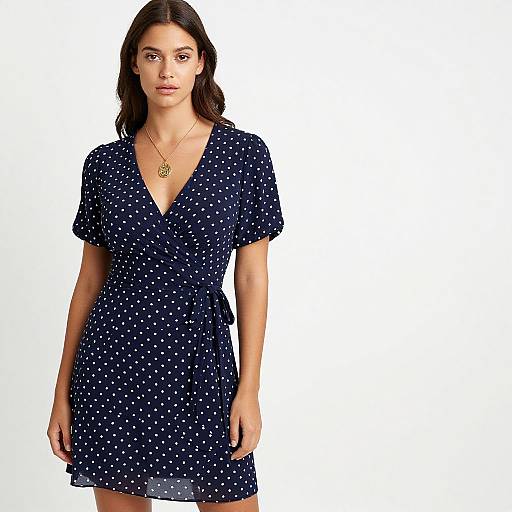 Photograph of a young woman with long dark hair, wearing a navy blue polka dot dress with short sleeves and a V-neck, and a gold