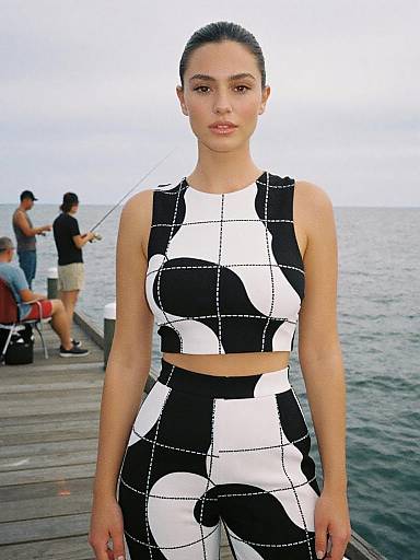 Photograph of a young woman with dark hair in a bun, wearing a black-and-white geometric crop top and high-waisted pants, standing on