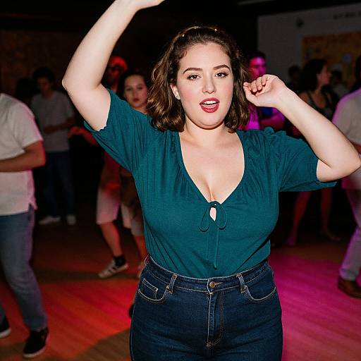 Photograph of a curvy, fair-skinned woman with short brown hair, wearing a teal blouse and dark jeans, dancing energetically in a