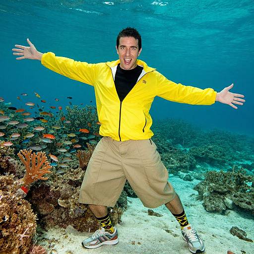 Photograph of a smiling man underwater, wearing a bright yellow jacket, beige cargo shorts, and white sneakers, with arms outstretched, surrounded by