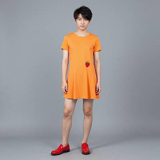 Photograph of an Asian man with short black hair, wearing an orange dress with a red strawberry graphic, and red shoes, standing against a plain gray