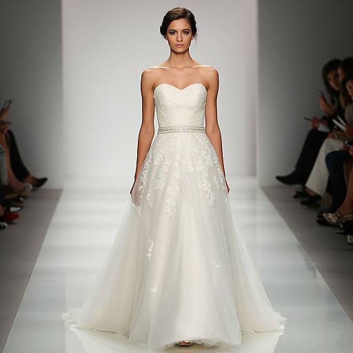 Photograph of a slender woman with dark hair in an elegant, strapless white lace wedding gown, walking down a brightly lit runway.