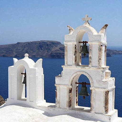Photograph of two white, weathered Greek Orthodox bell towers with crosses, overlooking a vivid blue sea and distant rocky island under a clear sky.
