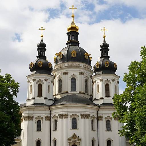 Photograph of a white Orthodox church with three black domes topped with golden crosses, set against a partly cloudy blue sky, flanked by green trees