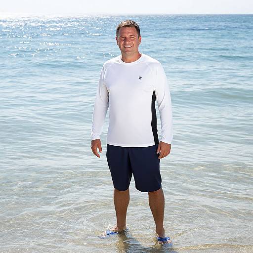 Photograph of a smiling man with short brown hair, wearing a white long-sleeve shirt and black shorts, standing in shallow, sunlit ocean