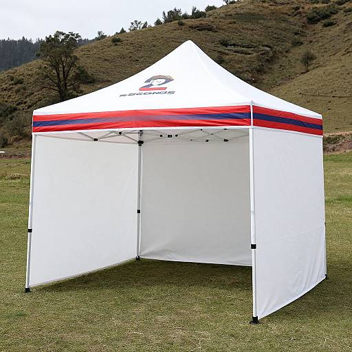 Photograph of a white, triangular pop-up tent with red and blue stripes on the peak, set on grassy field with hills and trees in the