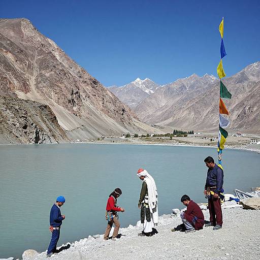 Photograph of five people near a mountain lake with snow-capped peaks, colorful prayer flags, wearing winter clothes, and standing/sitting on white gravel