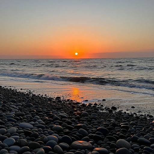 Photograph of a serene beach at sunset, featuring a pebble-covered shoreline, gentle waves, and a vibrant orange sun reflecting on the water.