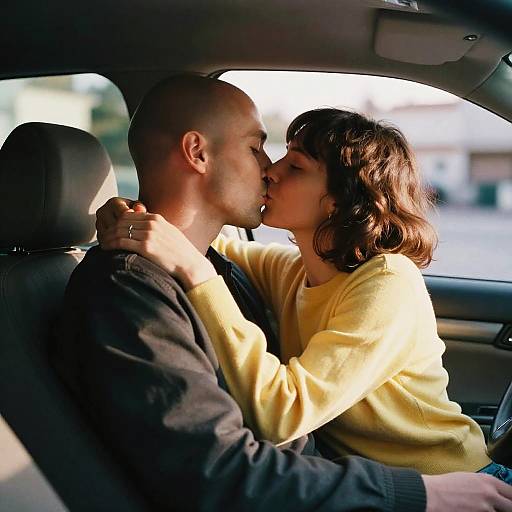 Photograph of a bald, light-skinned man and a brunette woman with shoulder-length hair kissing in a car, both wearing yellow and black sweaters