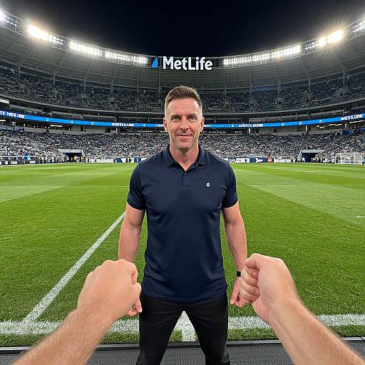 Photograph of a muscular, middle-aged man in a black polo shirt, standing on a soccer field, shaking hands, stadium full of spectators in background