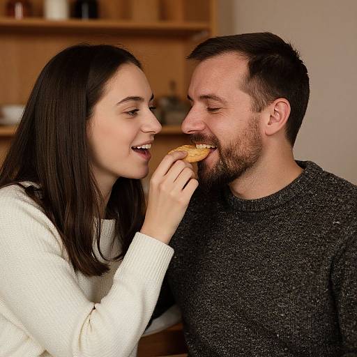 Photograph of a smiling couple sharing a donut in a cozy kitchen; woman with long dark hair in white sweater, man with short dark hair and