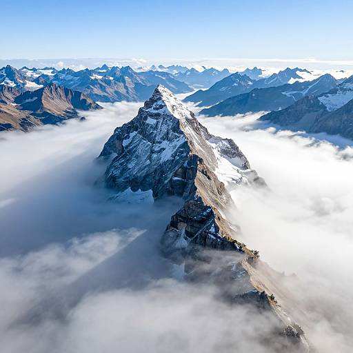 Aerial photograph of a snow-capped mountain peak surrounded by clouds, with a clear blue sky and distant mountain range in the background.