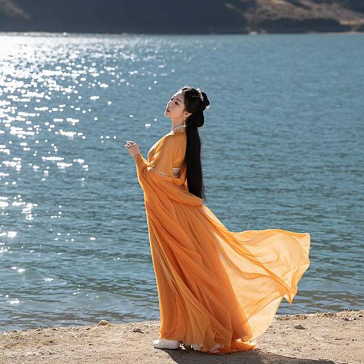 Photograph of an Asian woman with long black hair, wearing an orange flowing dress, standing by a sunlit, sparkling lake, looking up with a
