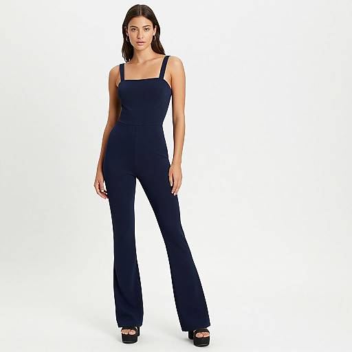 Photograph of a tall, slender woman with straight black hair wearing a black, sleeveless, square-shouldered jumpsuit and black high-heeled