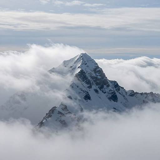 Photograph of a snow-capped mountain peak piercing through a layer of thick, white clouds, with a bright, partly cloudy sky above.