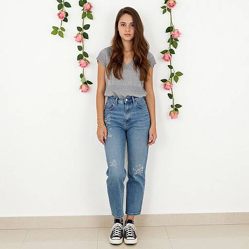 Photograph of a young woman with long brown hair, wearing a gray polka dot top and blue distressed jeans, standing against a white wall adorned with