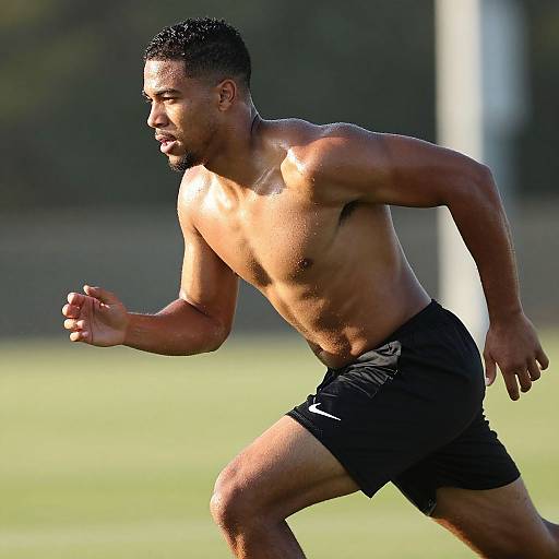 Photograph of a muscular, shirtless Black man with short curly hair, running on a grassy field in black shorts, sunlight highlighting his defined abs