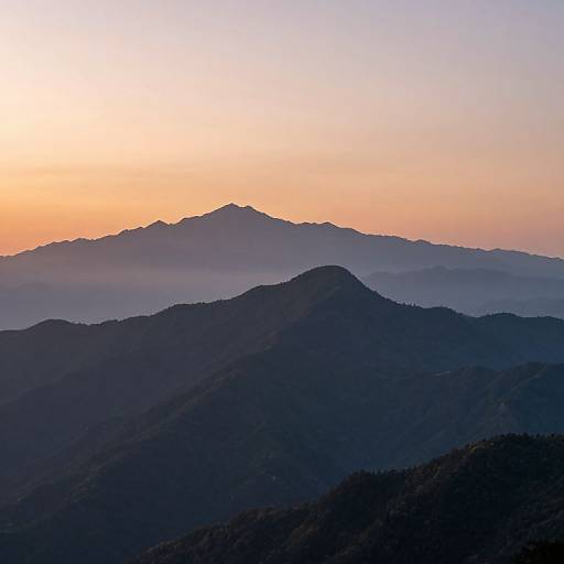 Photograph of a serene mountain landscape at sunset, featuring layered dark blue and purple mountains with a gradient orange and pink sky in the background.