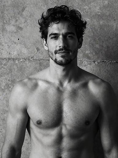 Black-and-white photograph of a muscular, shirtless man with curly hair and a trimmed beard, standing against a textured concrete wall.