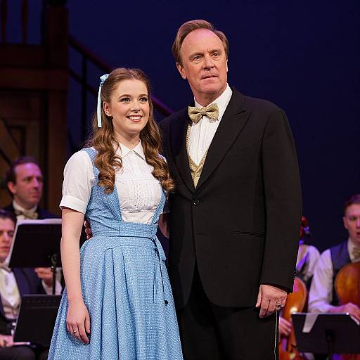 Photograph of a young woman with long brown hair in a blue dress and white blouse, standing beside an older man in a black suit and bow tie