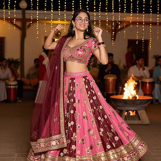 Photograph of a smiling Indian woman in a maroon and pink traditional lehenga with gold embroidery, dancing under string lights, in front of a burning