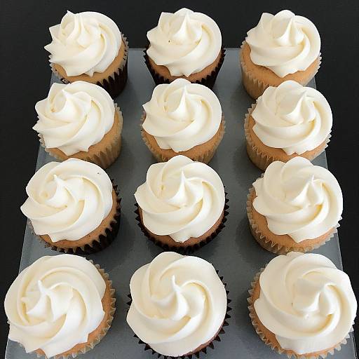 Photograph of twelve vanilla cupcakes with thick, white swirls of frosting, arranged in a 3x4 grid on a black tray.
