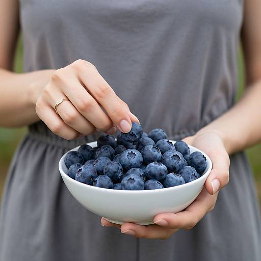 Photograph of a woman in a gray dress, holding a white bowl of fresh blueberries, using her right hand to pick one.