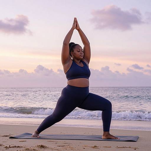 Photograph of a curvy Black woman in a navy sports bra and leggings, performing a yoga pose on a beach at sunset, with waves and a